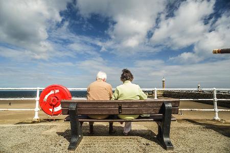 Ein altes Ehepaar sitzt auf einer Bank am Hafen mit Blick zu...