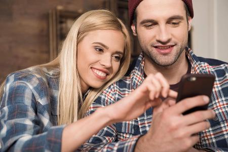 Smiling young couple using smartphone together at home model...