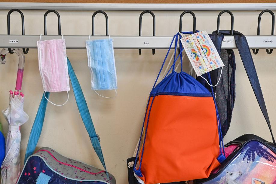 Masken hängen zusammen mit Taschen und Rucksäcken an Kleiderhaken in einem Klassenraum einer Grundschule.