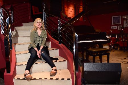 Frau sitzt auf Treppe und lächelt in die Kamera, während im Hintergrund verschiedene Musikinstrumente zu sehen sind.