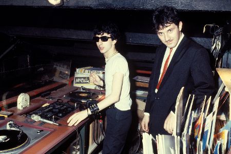 Zwei Männer stehen hinter einem veralteten DJ-Pult, neben ihnen liegen mehrere Schallplatten.