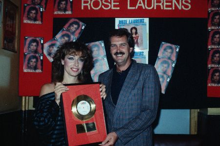 Frau mit Goldener Schallplatte in der Hand und Mann posieren vor einer Wand mit Postern.