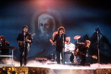 Fünf Männer stehen auf einer Bühne und performen einen Song, teilweise mit Instrumenten
