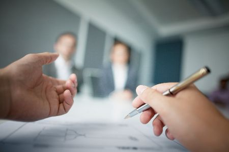 Hände mit Stift gestikulieren über Papier, im Hintergrund zwei schemenhafte Personen.