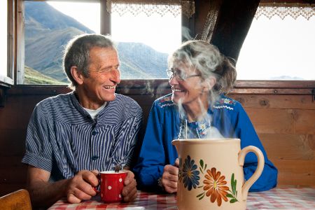 Zwei alte Menschen sitzen in einem Haus vor Bergpanorama und trinken Kaffee.