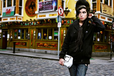 Junge Frau mit schwarzer Turmfrisur steht auf der Straße vor einem Pub, in der Hand eine Handtasche