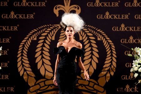 Frau mit weißer Hutkreation vor einem schwarz-goldenem Logo auf Laufsteg