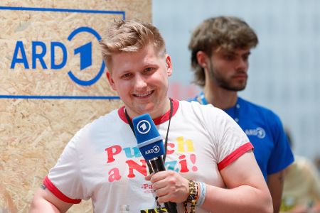 Mann mit Mikrofon in der Hand vor ARD-Logo
