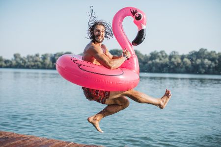 Mann mit pinkem Flamingo-Schwimmring springt vor Gewässer lachend in die Luft Mann mit pinkem Flamingo-Schwimmring springt vor Gewässer lachend in die Luft