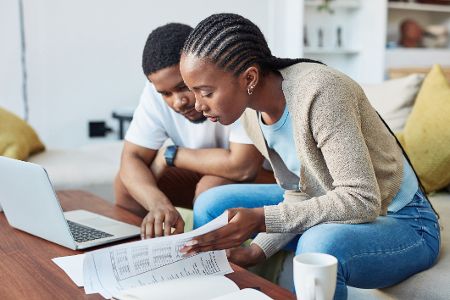 Mann und Frau sitzen vor einem Laptop und schauen in Papiere mit Zahlen drauf