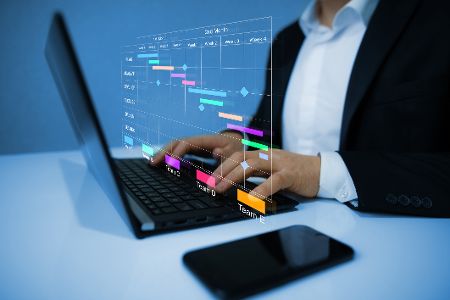 Mensch sitzt am Laptop und arbeitet mit dem Handy neben sich an Plänen