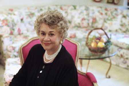 Joan Plowright sitzt auf einem Stuhl, im Hintergrund ist ein Plüschsofa zu sehen.