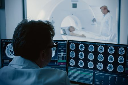 Im Vordergrund überwacht ein Arzt Daten, im Hintergrund wird ein Mensch in eine Röhre geschoben