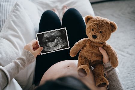 Schwangere Frau sitzt mit Teddybär und Ultraschallbild in der Hand auf dem Sofa