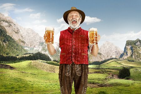 Älterer Mann in bayerischer Tracht mit zwei Maßkrügen mit Bier in der Hand posiert vor Bergkulisse
