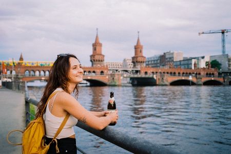 Frau lehnt an Geländer vor Berliner Brücke und trinkt ein Bier