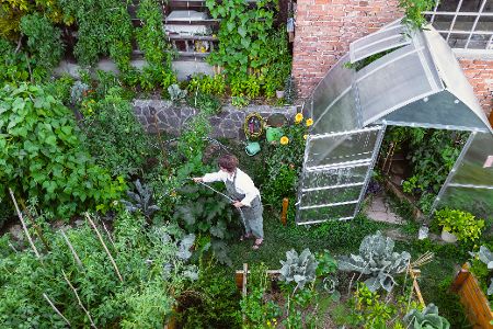 Kleiner Garten mit Gewächshaus wird von einer Person gewässert