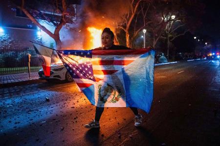 Eine Frau mit mehreren Flaggen geht an einem brennenden Auto vorbei, während sie gegen die Razzien der Trump-Regierung gegen Einwanderer in Los Angeles protestiert.