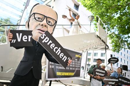Ein Aktivist mit Merz-Maske zerbricht während einer symbolischen Protestaktion von Kabul Luftbrücke und Campact ein Schild mit der Aufschrift 