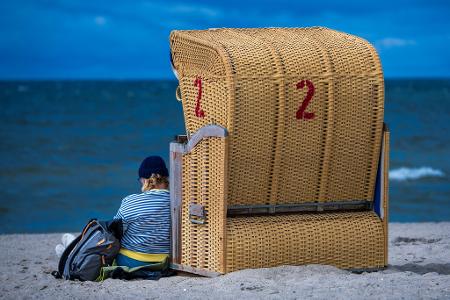 Regenschauer und kräftiger Wind an der Ostseeküste: Eine Frau sitzt mit Mütze bei wechselhaftem Wetter neben einem Strandkorb auf der Insel Poel.