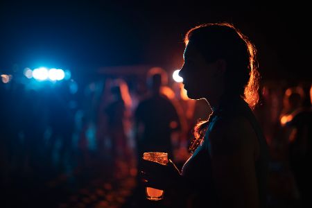 Frau mit einem Glas in der Hand steht draußen im Dunkeln, während im Hintergrund gefeiert wird