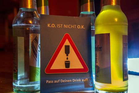 Ein Schild mit dem Hinweis, dass man auf sein Glas aufpassen soll, steht vor zwei Flaschen mit Alkohol