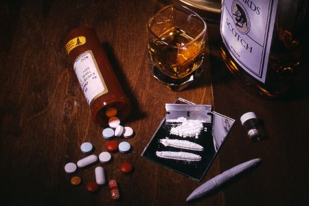 Flasche und Glas mit Alkohol neben verschiedenen anderen Drogen, darunter ein Joint, Tabletten und Kokain