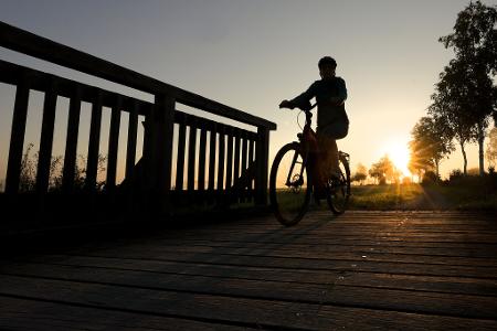 Eine Radfahrerin fährt in Bad Buchau im Sonnenaufgang über eine Brücke.