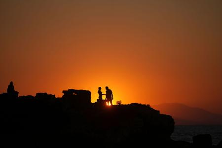 Ein Paar steht auf einem Meeresfelsen, während die Sonne an einem warmen und windigen Tag am Strand von Kavouri im Süden Athens untergeht,
