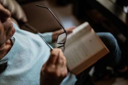Alte Frau mit Buch auf dem Schoß hat in der einen Hand eine Lesebrille