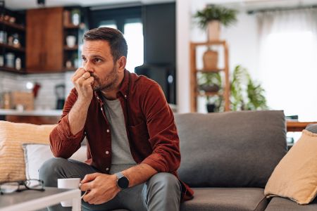 Nervöser Mann mit Kaffeetasse in der Hand sitzt an einem Couchtisch auf einem Sofa