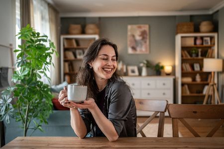 Glückliche Frau sitzt an einem Tisch in einer Wohnung und hat eine Tasse Kaffee in der Hand