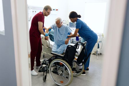 Zwei Pflegende helfen einer Patientin in den Rollstuhl