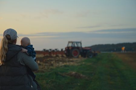 Mutter steht mit Baby auf dem Arm neben einem Feld auf dem Land, das gerade mit einem Traktor bearbeitet wird
