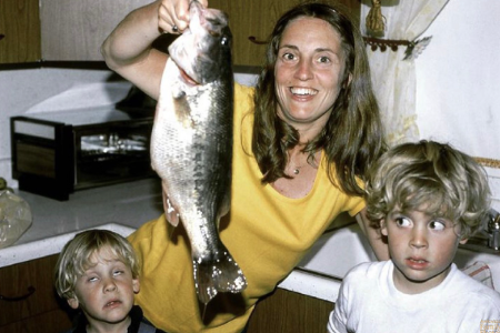 Frau hält Fisch in Kamera, während zwei Kinder die Augen verdrehen