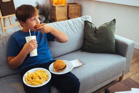 Kind sitzt mit Burger, Pommes und Softgetränk auf einer Couch