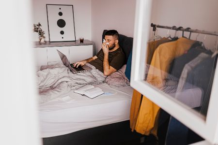 Mann liegt mit Laptop im Bett und putzt sich die Nase