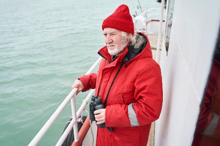 Mann mit Fernglas steht an der Reling eines Schiffes und schaut in die Ferne