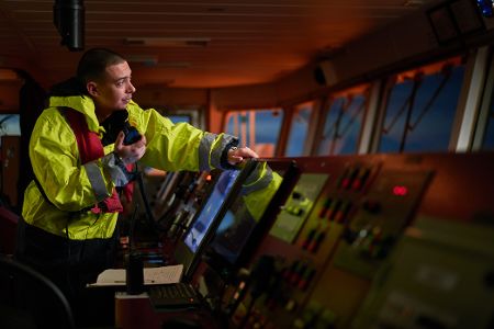 Mann in gelber Arbeitsjacke steht auf der Brücke eines Schiffes und funkt