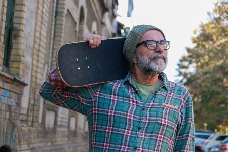 Älterer Mann mit Vollbart und Mütze hat ein Skateboard lässig auf der Schulter