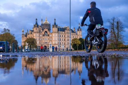 Herbstwetter in Norddeutschland: Ein Radfahrer fährt an einer Pfütze vorbei, in der sich das Schweriner Schloss spiegelt.