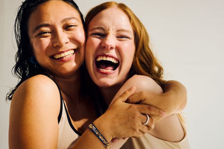 Zwei junge Frauen haben sich im Arm und lachen herzhaft in die Kamera