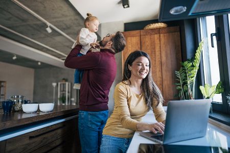 Mann spielt mit dem Baby in der Küche, während die Frau am Laptop arbeitet