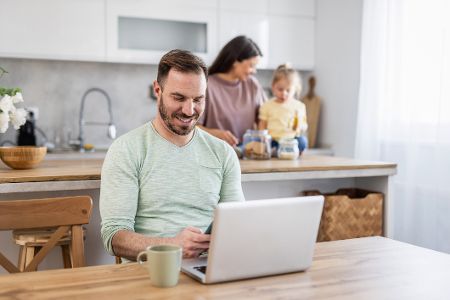 Frau steht mit Kind in der Küche, während der Mann am Laptop arbeitet