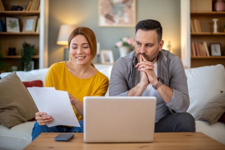 Frau und Mann sitzen im Wohnzimmer auf der Couch mit Papieren vor einem Laptop